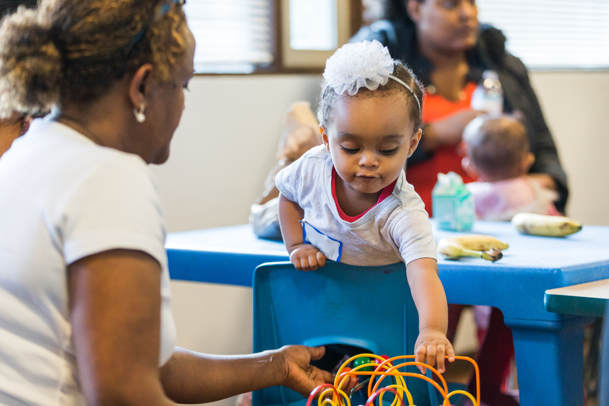 small child laying on table and leaning over a chair for a toy while a woman assists.