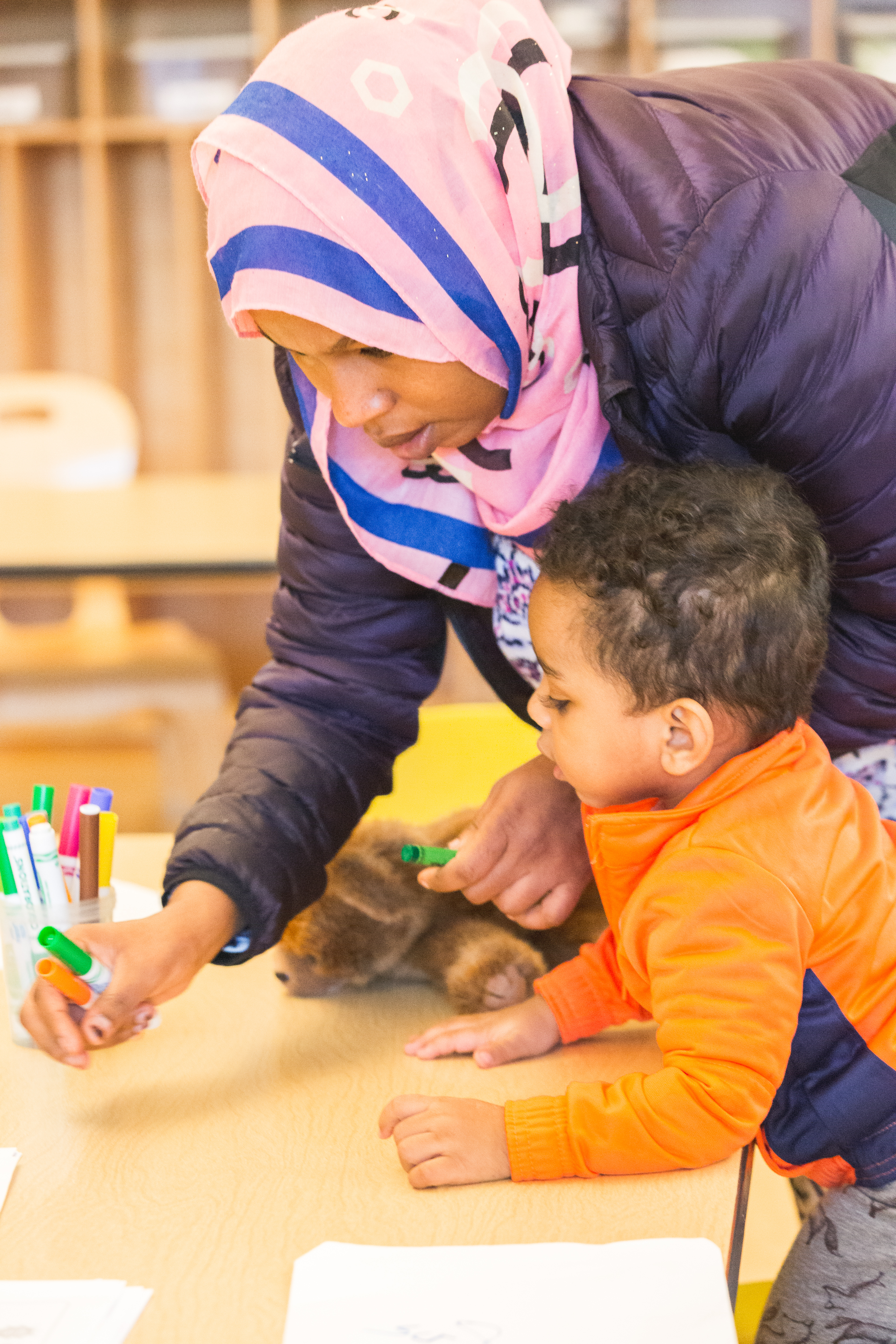 woman leaning over a toddler with a market and a sheet of paper they are looking at together