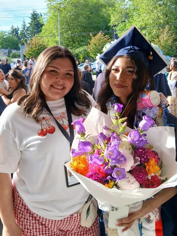 A young woman in a blue graduation cap and gown holding flowers next to another woman smiling