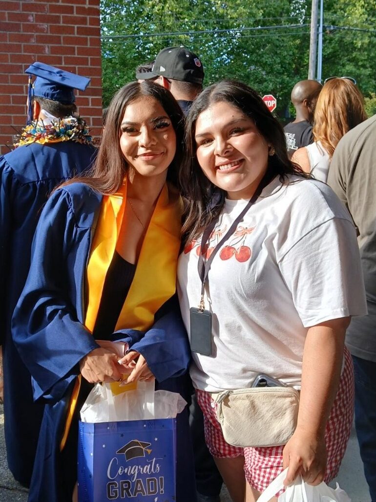 A young woman in a blue graduation gown holding a gift bag next to another woman smiling