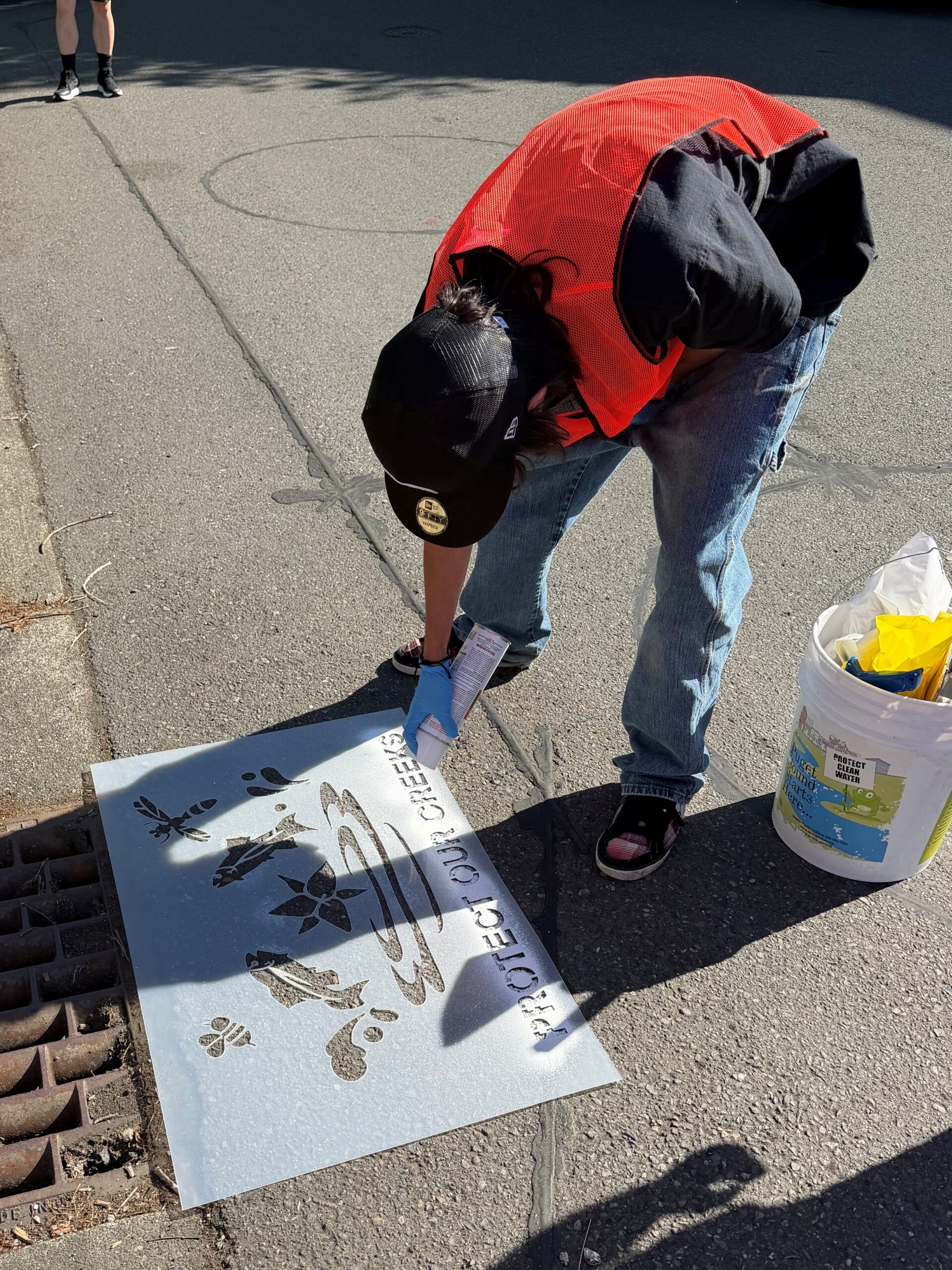 Man in jeans, a red shirt, and a black hat leaning over a stencil and spray panting the floor.