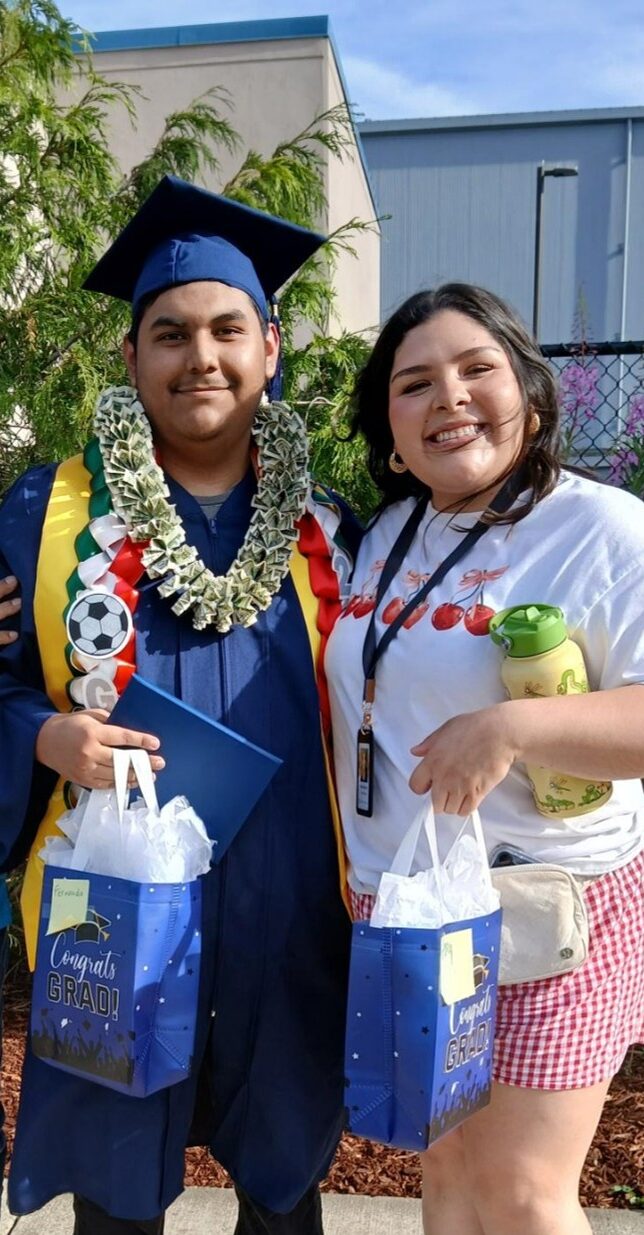 Young man holding a gift bag dressed in a blue graduation cap and gown smiling next to a woman holding a gift bag smiling.
