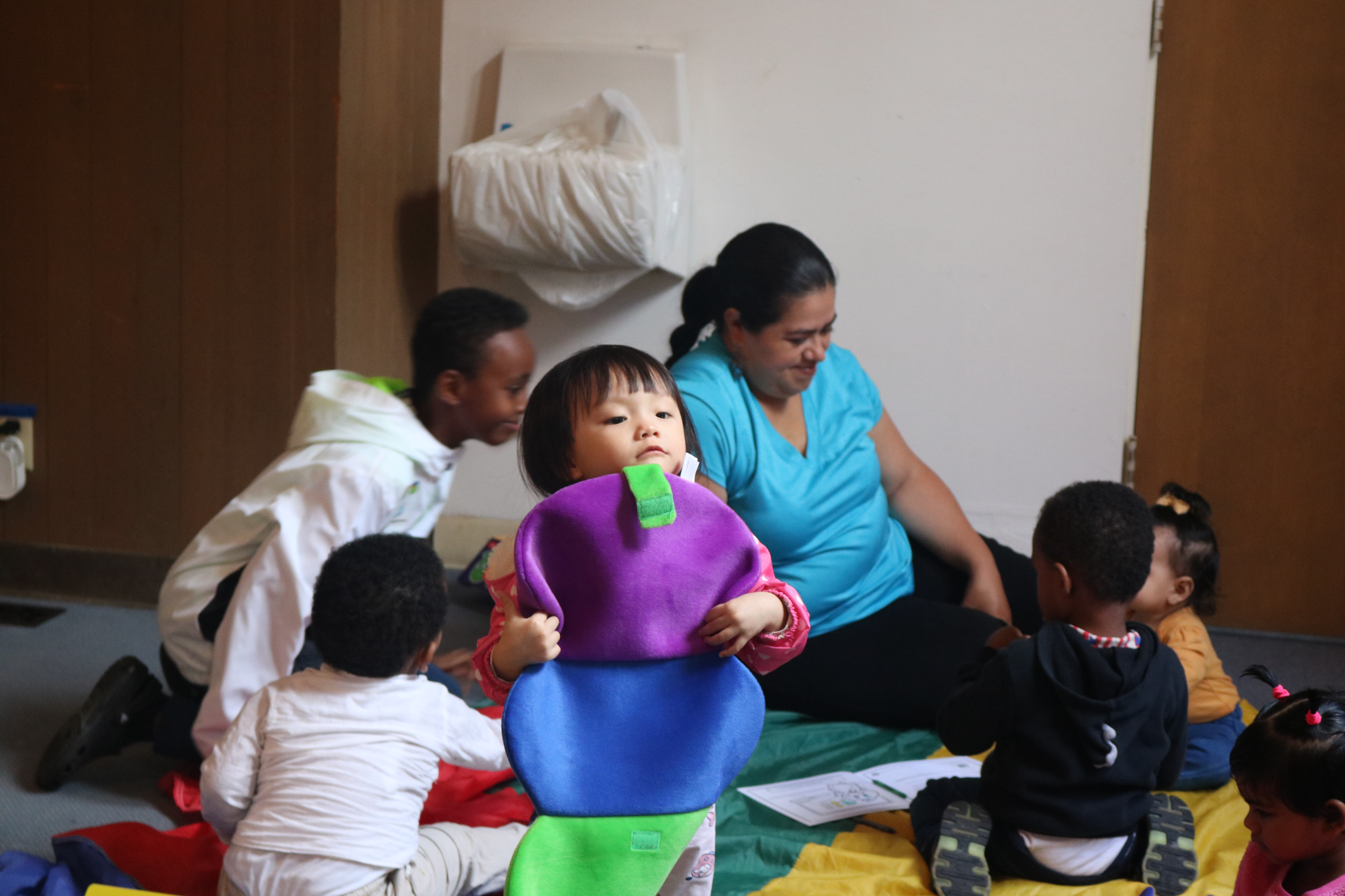 Small child playing with caterpillar felt toy with woman and toddlers in the background
