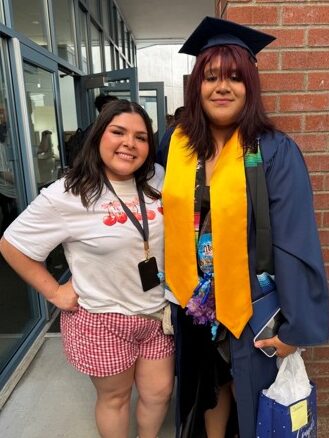 A young woman in a blue graduation cap and gown holding a gift bag next to another woman smiling