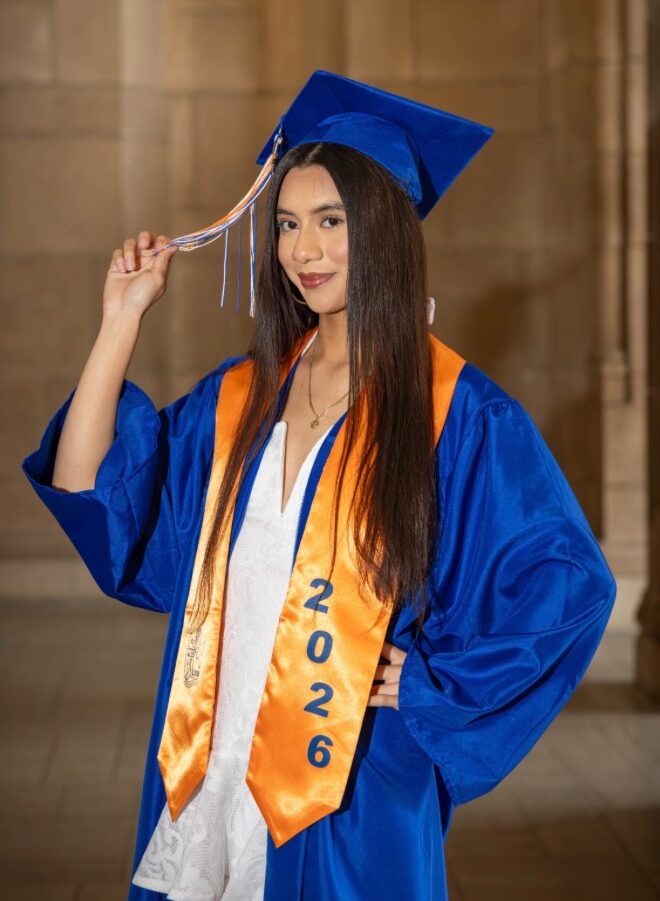 Young woman in a blue graduation cap and gown holding her tassle with a gold sash with a blue 2026 on it